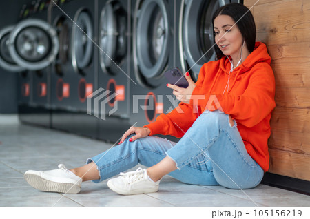 Young woman listening music with smartphone and earphones waiting in laundry room. Young woman listening music with smartphone and earphones waiting in laundry room. 105156219