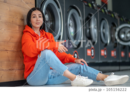 Young woman listening music with smartphone and earphones waiting in laundry room. 105156222