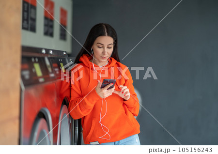 Young woman listening music with smartphone and earphones waiting in laundry room. 105156243