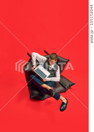 Businessman, employee sitting on armchair in weird, uncomfortable position, working on laptop against red studio background 105158044