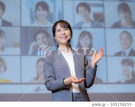 ホールで観衆とテレビ電話でつながっている人物を相手に講演をする女性講師 105158255