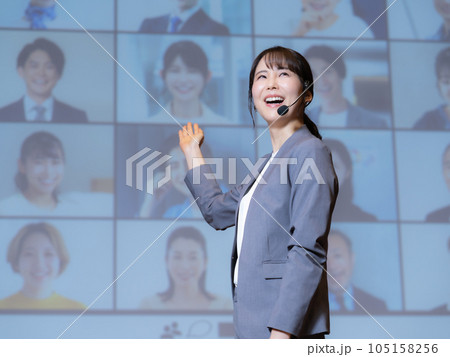 ホールで観衆とテレビ電話でつながっている人物を相手に講演をする女性講師 105158256