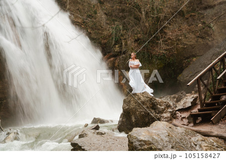 Happy woman in a white dress stands on a stone with a waterfall behind Happy woman in a white dress stands on a stone with a waterfall behind 105158427