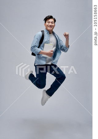 Full length of a smiling young asian man carrying backpack isolated, holding books, jumping Full length of a smiling young asian man carrying backpack isolated, holding books, jumping 105158663