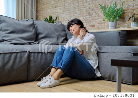 Mature woman suffering from neck pain, sitting on floor in living room 105159601