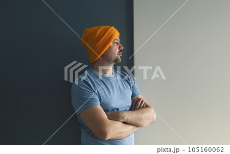 Young man in a blue T-shirt and a yellow hat stands with his arms crossed against a gray-blue wall. 105160062