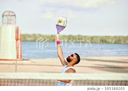 Dynamic image of bearded man playing beach, paddle tennis, hitting ball with racket. Outdoor training on warm summer day Dynamic image of bearded man playing beach, paddle tennis, hitting ball with racket. Outdoor training on warm summer day 105160203