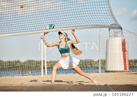 Competitive woman, athlete, professional tennis player in motion during game, hitting ball with racket, training on beach on summer day 105160227