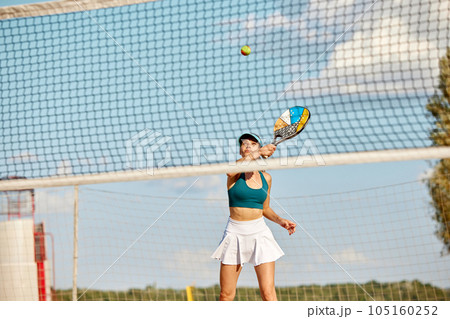 Dynamic image of young woman playing beach tennis, hitting ball with racket. Outdoor training on warm summer day 105160252
