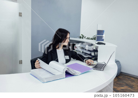 Office female worker in black jacket and white shirt works with catalog of papers and laptop Office female worker in black jacket and white shirt works with catalog of papers and laptop 105160473