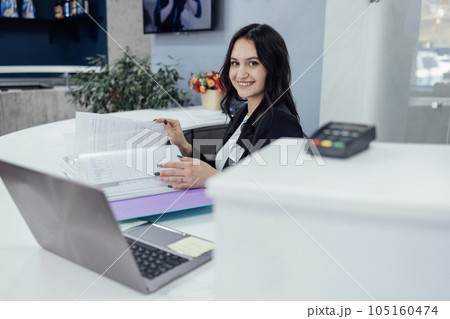 Office female worker in black jacket and white shirt works with catalog of papers and laptop Office female worker in black jacket and white shirt works with catalog of papers and laptop 105160474