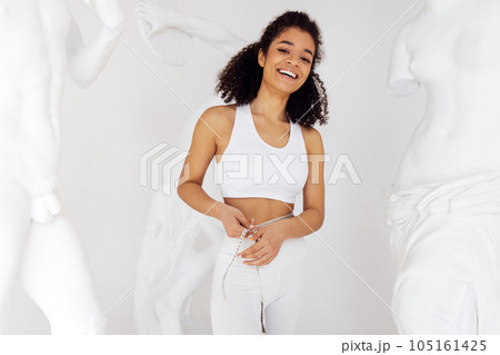 Slender attractive african woman in white sportswear measuring waist using tape. Slender attractive african woman in white sportswear measuring waist using tape. 105161425