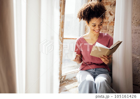 Young smiling pretty mixed race girl sits on windowsill with porcelain cup of tea and book. 105161426