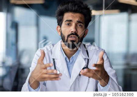 Online medical consultation. Portrait of a serious young Indian male doctor sitting in the office at the table talking, consulting the camera, gesturing with his hands. Online medical consultation. Portrait of a serious young Indian male doctor sitting in the office at the table talking, consulting the camera, gesturing with his hands. 105162300