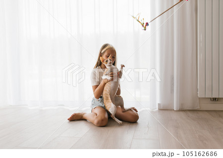 Caucasian teen girl plays with cornish rex cat at home on a sunny day, hugs him and laughs while sitting on the floor in the living room 105162686