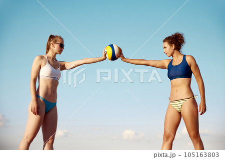 Two sportive young womsn, with fit, slim body in swimsuit holding volleyball ball over blue sky background Two sportive young womsn, with fit, slim body in swimsuit holding volleyball ball over blue sky background 105163803