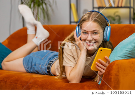 Happy young woman in wireless headphones relaxing lying on sofa at home listening favorite music 105168011