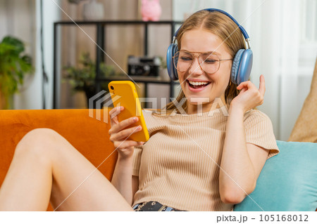 Happy young woman in wireless headphones relaxing lying on sofa at home listening favorite music 105168012