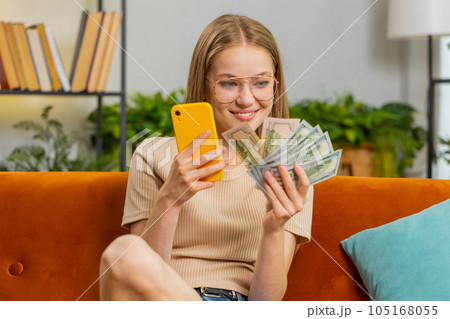 Smiling happy young woman counting money cash use smartphone income saves lottery win budget at home Smiling happy young woman counting money cash use smartphone income saves lottery win budget at home 105168055