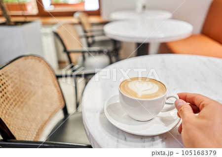 A man holds a cup with his hand A cup of hot cappuccino on the table in the coworking space of the coffee shop. Coffee break at the cafe. Close-up A man holds a cup with his hand A cup of hot cappuccino on the table in the coworking space of the coffee shop. Coffee break at the cafe. Close-up 105168379