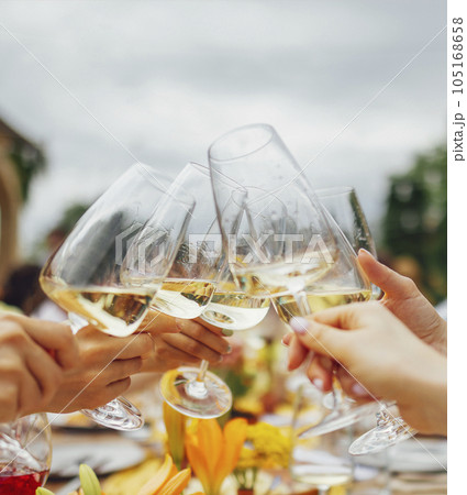 Female hands holding elegant glasses with long stem of champagne and and clink them on the background of a table with snacks and flowers. 105168658