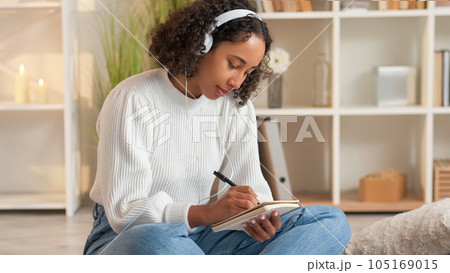 Goal achievement. Home rest. Dream planning. Inspired woman in headphones writing in diary sitting sofa in living room. 105169015
