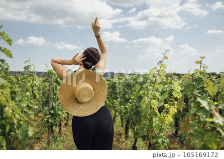 Young woman in black pants and wide brimmed straw hat stands in graceful pose and looks at vineyard. Young woman in black pants and wide brimmed straw hat stands in graceful pose and looks at vineyard. 105169172