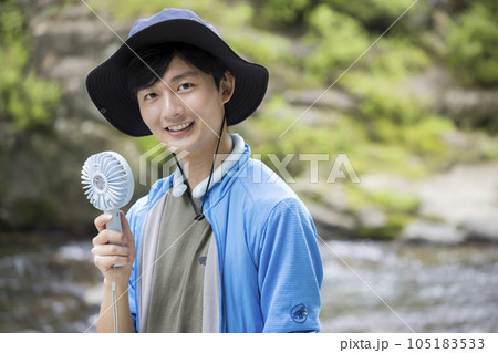 川辺でクールネックリングをして携帯扇風機を使う若い男性 105183533
