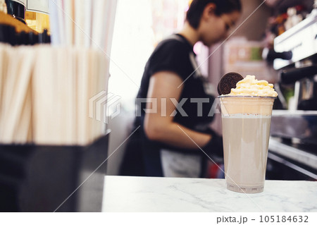 Young brunette female barista in black shirt, apron and black gloves decorates milk cocktail 105184632