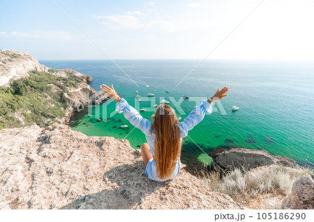 Woman travel sea. Happy woman in a beautiful location poses on a cliff high above the sea, with emerald waters and yachts in the background, while sharing her travel experiences Woman travel sea. Happy woman in a beautiful location poses on a cliff high above the sea, with emerald waters and yachts in the background, while sharing her travel experiences 105186290