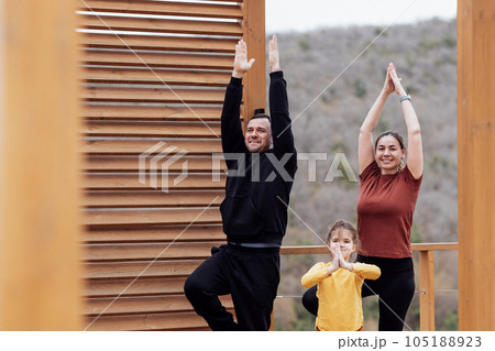 Happy family doing yoga outdoors. Attractive mom, handsome dad and cute daughter are standing in tree pose. Happy family doing yoga outdoors. Attractive mom, handsome dad and cute daughter are standing in tree pose. 105188923