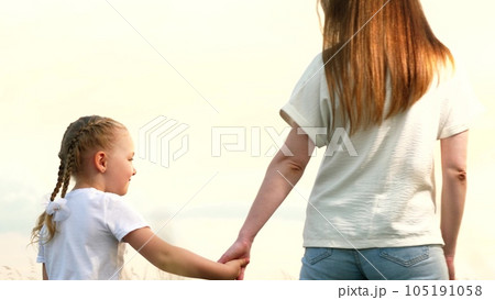 Mother and little daughter walk joining hands across field at sunset light backside view. Happy daughter holds mother hand walking together on country meadow. Mother and daughter enjoy family time 105191058