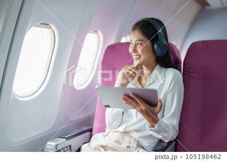 Young Asian attractive business woman passenger sitting on business class luxury plane while wearing wireless headphone for relax and working with tablet during the flight. 105198462