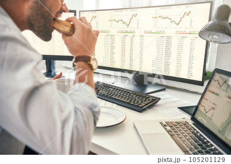 Enjoying lunch. Close-up of bearded trader is eating fresh sandwich and looking at monitor screen with financial data while sitting in his modern office Enjoying lunch. Close-up of bearded trader is eating fresh sandwich and looking at monitor screen with financial data while sitting in his modern office 105201129