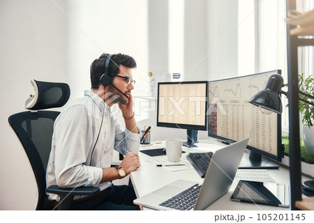 Always available. Side view of young bearded trader in headset talking with client and looking at monitor screen with trading charts and financial data while sitting in his modern office. Always available. Side view of young bearded trader in headset talking with client and looking at monitor screen with trading charts and financial data while sitting in his modern office. 105201145