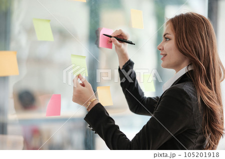 Beautiful female employee write down on colorful sticky notes manage list, concentrated biracial woman work on startup brainstorm collaborate plan on stickers on glass wall. Beautiful female employee write down on colorful sticky notes manage list, concentrated biracial woman work on startup brainstorm collaborate plan on stickers on glass wall. 105201918