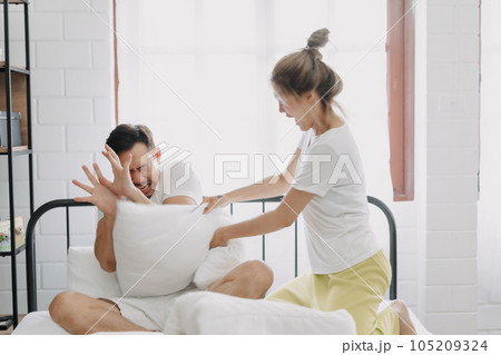 Funny couple angry wife hit husband with pillow in white bedroom. Funny couple angry wife hit husband with pillow in white bedroom. 105209324