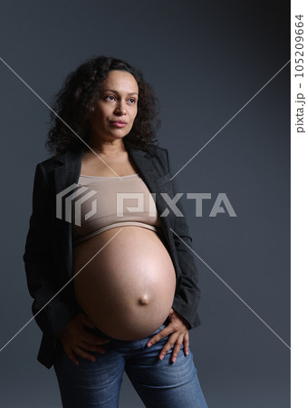 Curly haired pregnant woman with naker belly, wearing beige bra, gray blazer and blue jeans, thoughtfully looking aside Curly haired pregnant woman with naker belly, wearing beige bra, gray blazer and blue jeans, thoughtfully looking aside 105209664