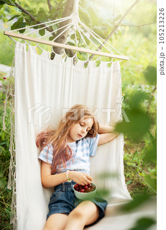 A little girl rests in a hammock and eats cherries in the summer. A little girl rests in a hammock and eats cherries in the summer. 105213226