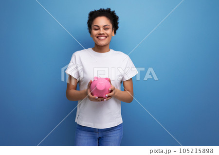 young happy brunette woman with afro hair in a white tank top holding a pink piggy bank. e-business young happy brunette woman with afro hair in a white tank top holding a pink piggy bank. e-business 105215898