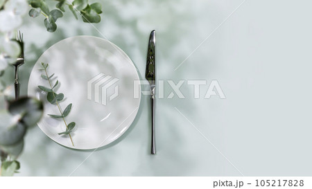 Spring holiday table settings. Empty plate with fork and knife on blue background with eucalyptus leaves. Mockup of romantic summer holiday card with setting suitable for menu. Flat lay, copy space 105217828