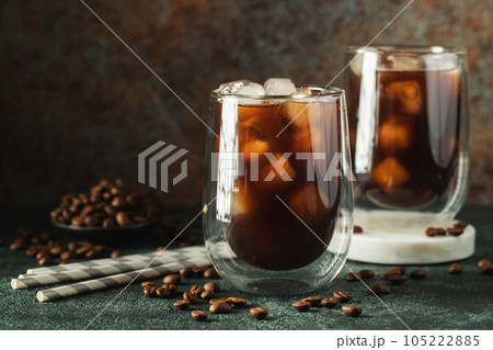 Ice coffee in a tall glass with cream poured over, ice cubes and beans on a dark concrete table. 105222885