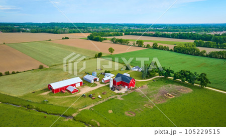 Green pastures and farmland around classic red barn with tractors and distant dirt fields aerial Green pastures and farmland around classic red barn with tractors and distant dirt fields aerial 105229515