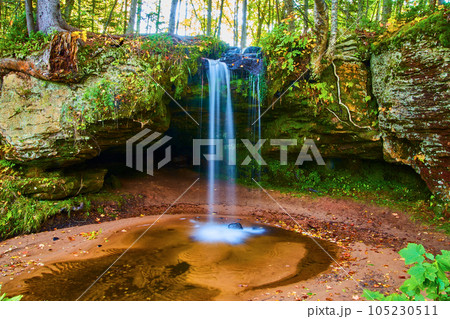 Tall blue waterfall pouring over lichen, moss covered cliff edge into shallow basin in summer forest 105230511