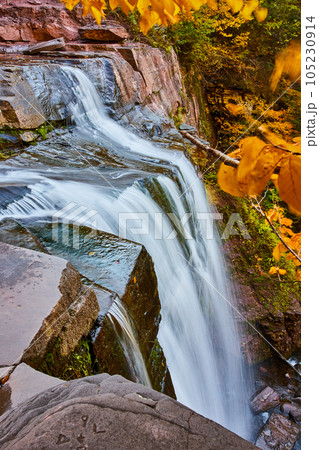 On edge of rocky cliff by huge waterfall pouring over edge with yellow leaves around 105230914