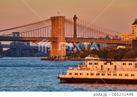 Governors Island Ferry in golden hour sunset dusk light from waters passing by Brooklyn Bridge with American Flag Governors Island Ferry in golden hour sunset dusk light from waters passing by Brooklyn Bridge with American Flag 105231446