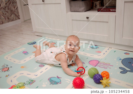 A cute baby in a white suit sits on a soft mat and plays with bright toys. High quality photo 105234458