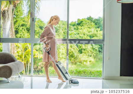 Young pregnant woman enjoys cleaning her house. Easy cleaning with a wireless vacuum cleaner. Pregnant woman cleaning floor with handheld vacuum cleaner. Modern easy cleaning 105237516