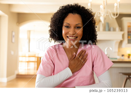 Happy biracial female medical worker wearing scrubs having video call in kitchen at home, listening 105239313