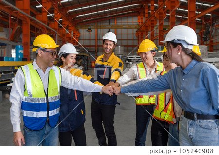 Group of construction workers clad in hard hats and reflective vests, are locked in a cordial fist bump, signaling joint progress. Machinery and steel beams compose the factory backdrop. Group of construction workers clad in hard hats and reflective vests, are locked in a cordial fist bump, signaling joint progress. Machinery and steel beams compose the factory backdrop. 105240908